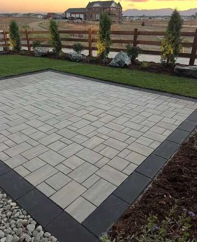 Pale rectangular paver patio with a dark perimeter border and sunset beyond the fence.