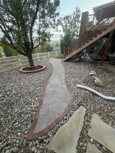 Curved textured walkway with reddish edging winding through river rock beside a deck.