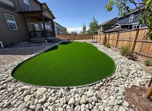Curved artificial turf lawn edged with smooth river rock beside a home.