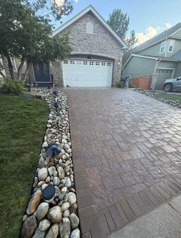 Decorative patterned driveway bordered by river rock and landscape lights.